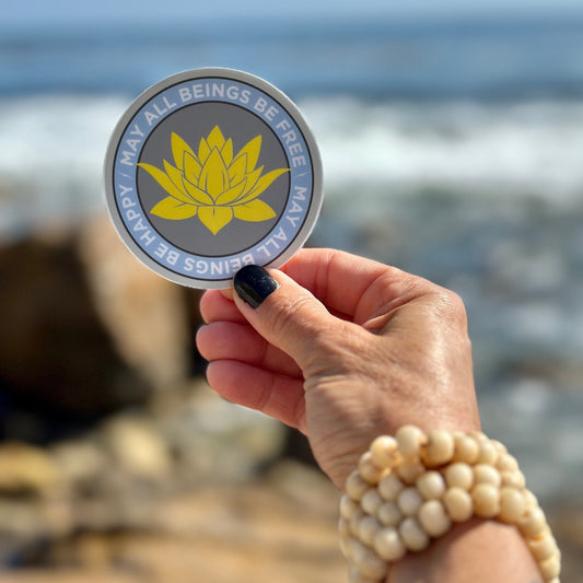 Hand holding a 3" circular sticker with a lotus flower design and text, may all beings be free may all beings be happy, against a blurred beach background.