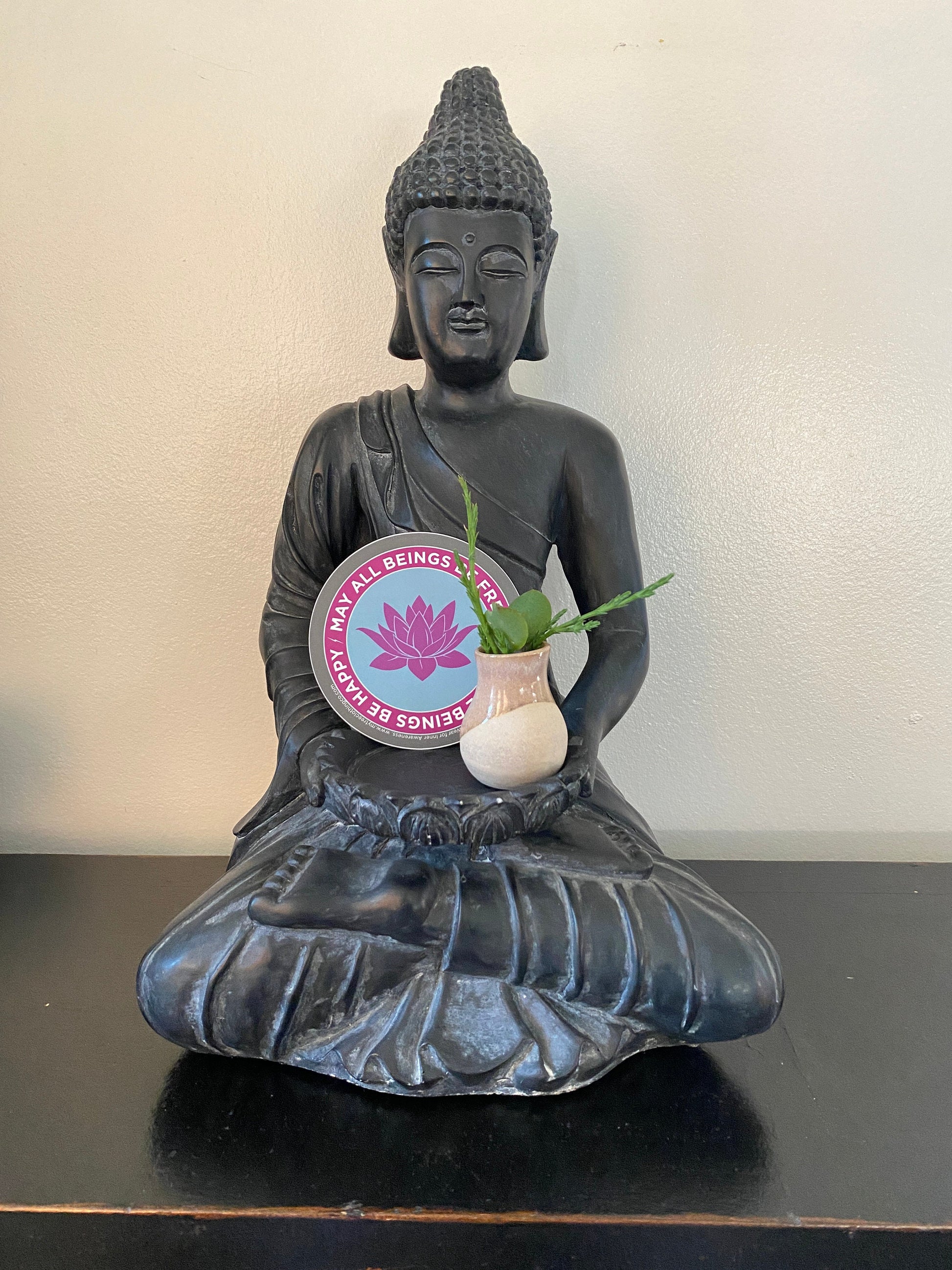Buddha statue holding a small plant with a 4" circular Buddhist sticker with a purple lotus flower and text, may all beings be free may all beings be happy.