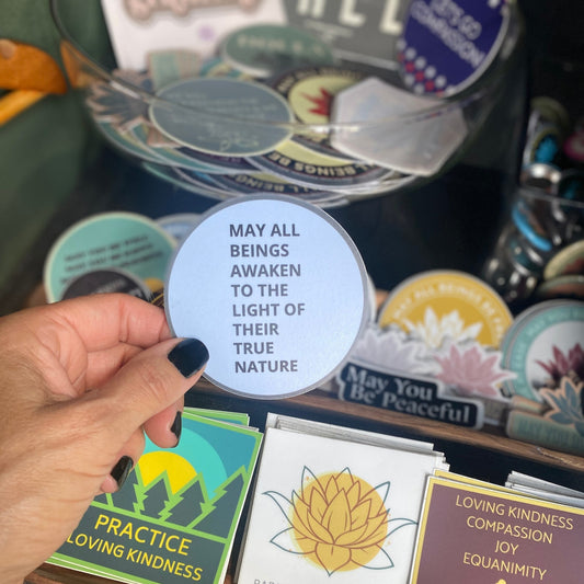 Hand holding a circular sticker with the text, may all beings awaken to the light of their true nature, in front of a display of other stickers.