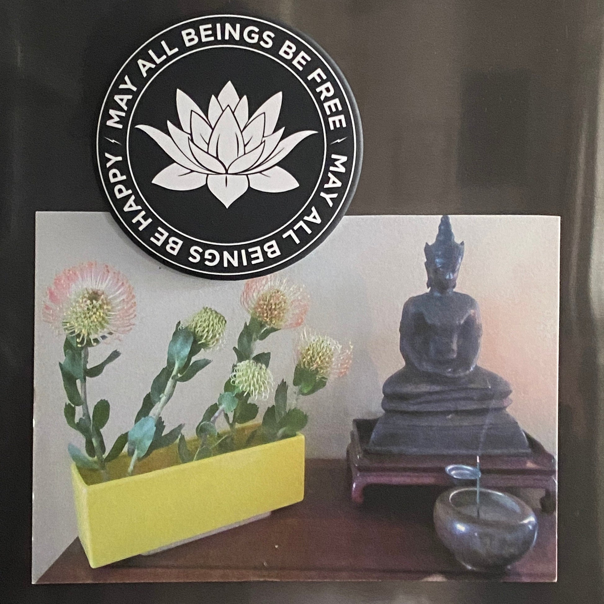 Decorative card with a Buddha statue, flowers, attached to a refrigerator by a 3.5" round black and white magnet with Buddhist lotus flower design and text, may all beings be free, may all beings be happy.