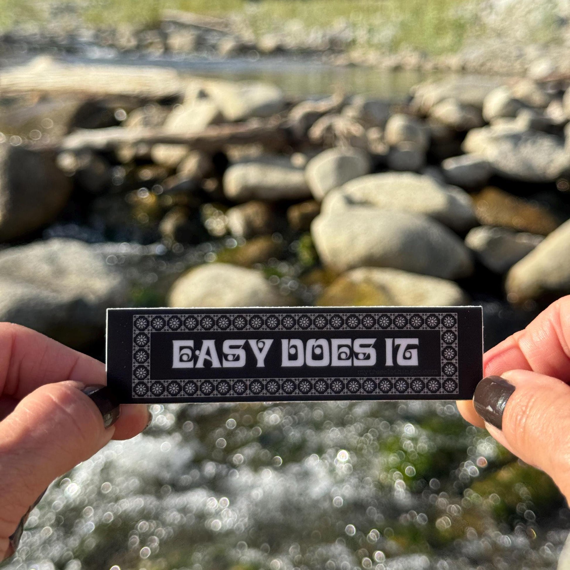Person holding a 'Easy Does It' sticker with a natural background of rocks and water.