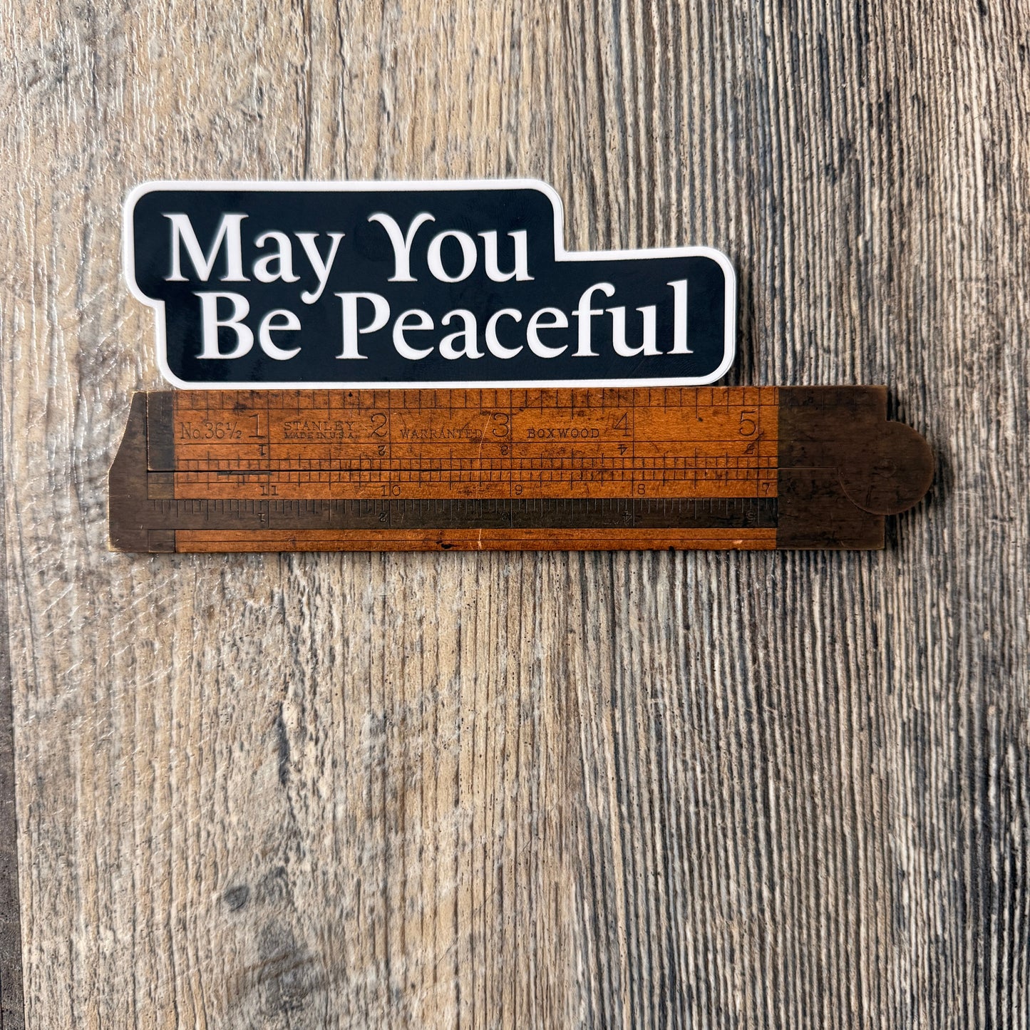 Five inch by one point five inch black and white die cut sticker that reads, MAY YOU BE PEACEFUL'. It's being measured by an antique wooden ruler.