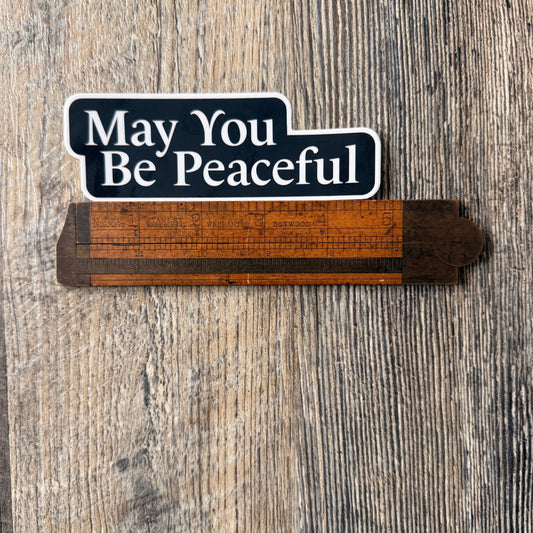 Five inch by one point five inch black and white die cut sticker that reads, MAY YOU BE PEACEFUL'. It's being measured by an antique wooden ruler.
