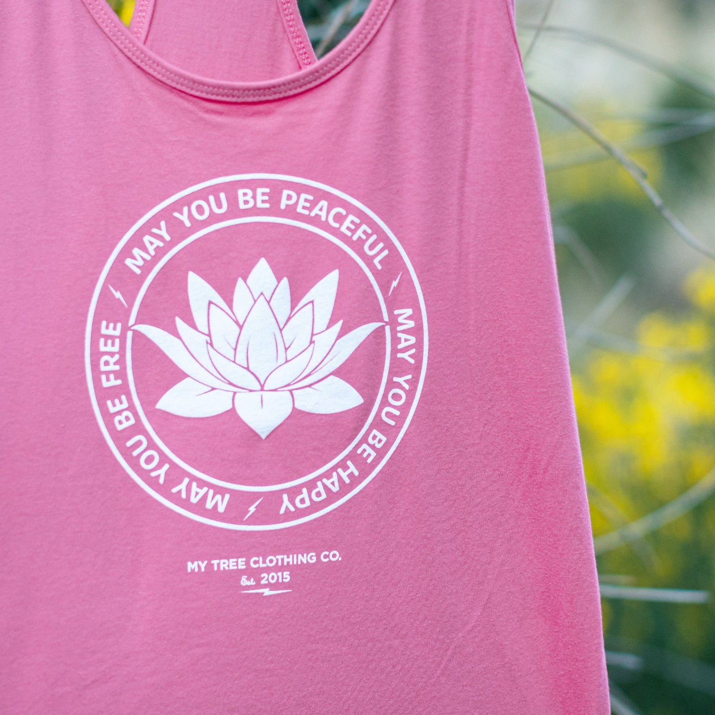 Close up photo of a pink tank top with a white lotus flower design and text that reads, 'may you be peaceful may you be happy may you be free.'
