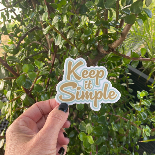 Hand holding a 12 Step recovery slogan sticker in gold and blue with the text, 'Keep it Simple' in front of green foliage.