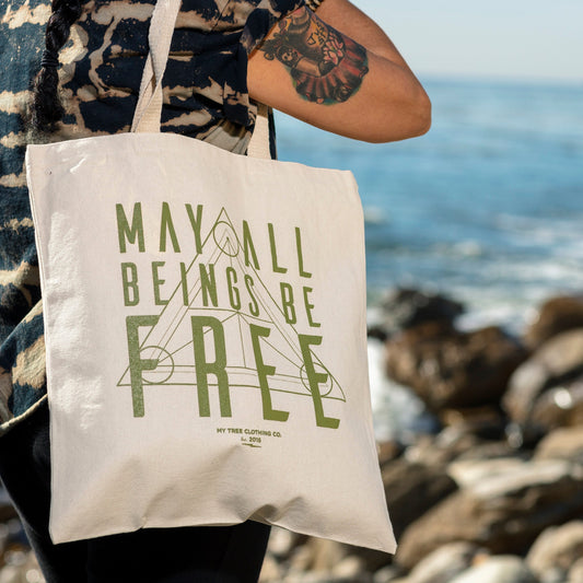 Person holding a tote bag with Buddhist mantra, 'May All Beings Be Free' text by the ocean.
