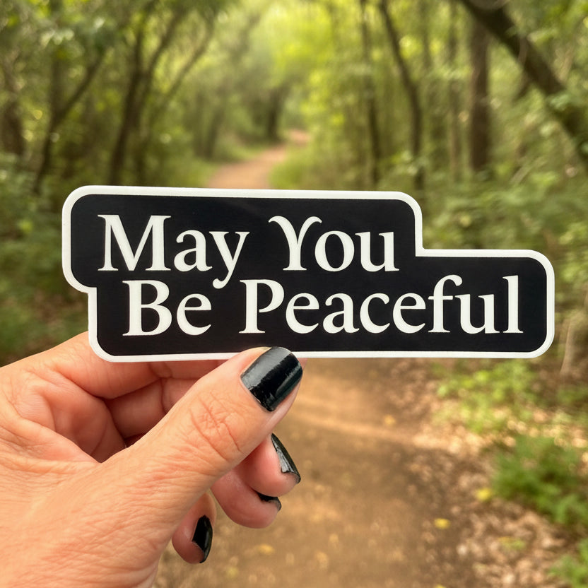 Five inch by one point five inch black and white die cut sticker that reads, MAY YOU BE PEACEFUL'. It is being held in a hand with a nature trail in the background.