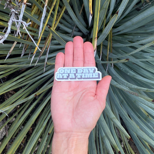 Hand holding a 'One Day at a Time' sticker in front of a plant.
