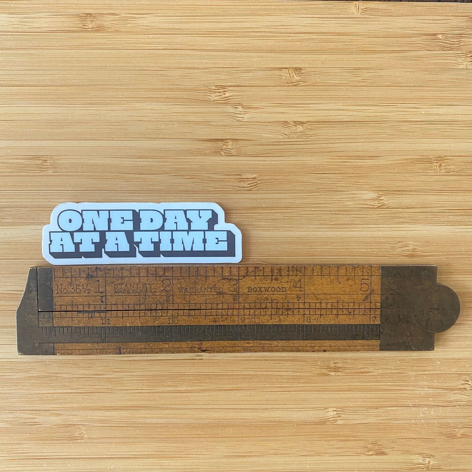 Sticker with 'ONE DAY AT A TIME' text on a wooden surface with a ruler.