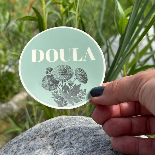 Round green sticker with 'DOULA' text held by a hand against a natural background.