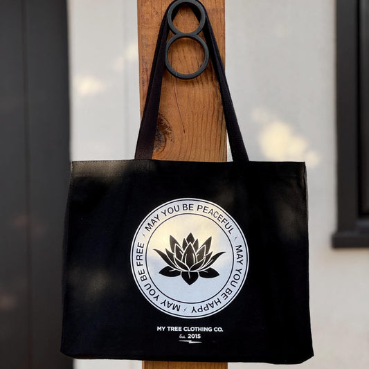 Black tote bag with a circular design featuring a lotus flower and text, may you be peaceful may you be happy may you be free, hanging on a wooden door.