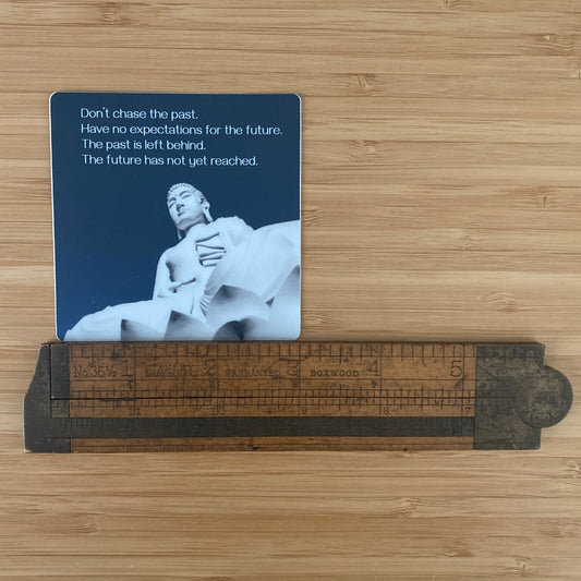 3" round sticker with a Buddha statue and text, "don't chase the past, have no expectations for the future, the past is left behind, the future has not yet reached" it is being measured by an antique ruler.