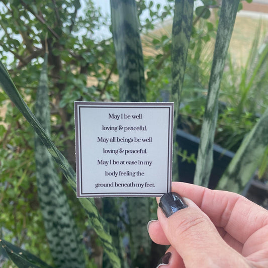 Hand holding a 2" square Buddhist prayer in front of plants.