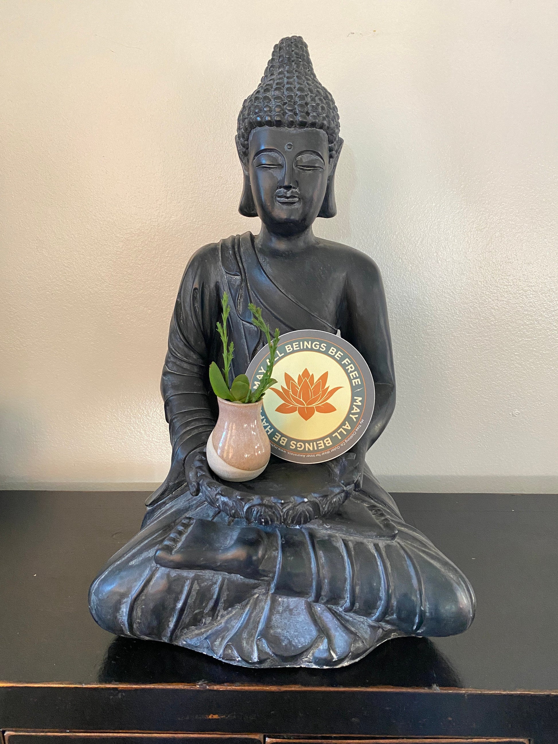Buddha statue holding a pot with a plant on a plain background with a 4" circular sticker with a saffron colored lotus flower design and Mettā prayer.