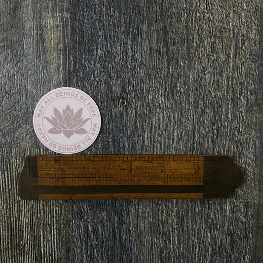 Wooden ruler with measurement markings on a textured gray surface, featuring a 2" circular pink Buddhist lotus flower sticker that reads, may all beings be free may all beings be happy.
