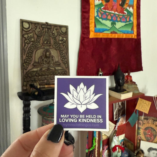 Hand holding a 2" square purple and white  lotus sticker with the words, may you be held in loving kindness.