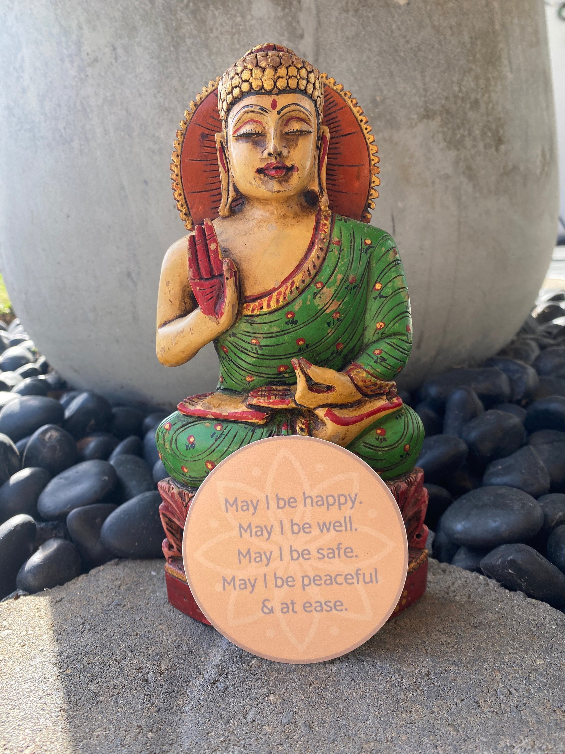 Colorful Buddha statue with a circular sticker with text, may i be happy may i be well may i be safe may i be peaceful may i be at ease, on pebbles.