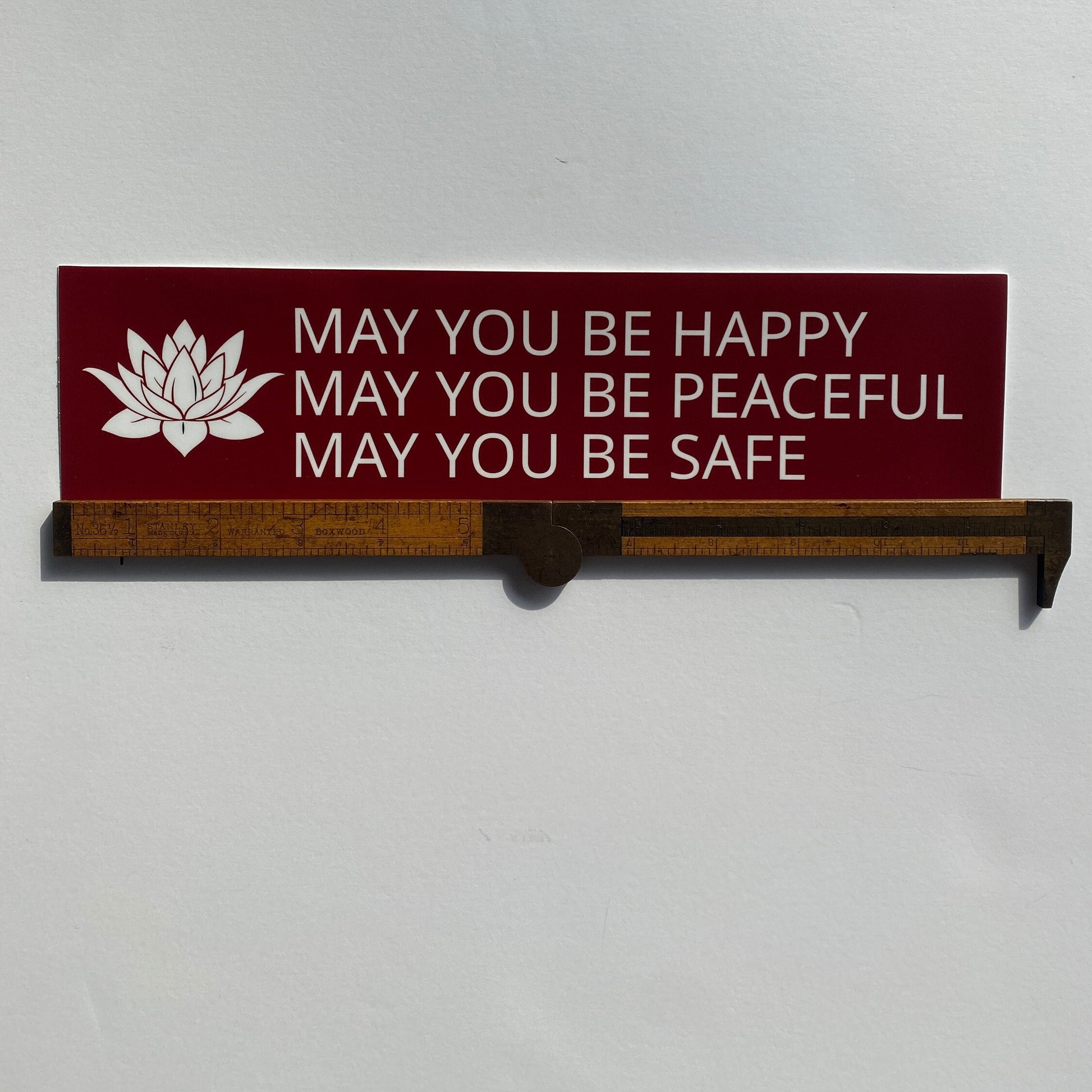 Red and white bumper sticker with a lotus and the prayer, may you be happy may you be peaceful may you be safe.