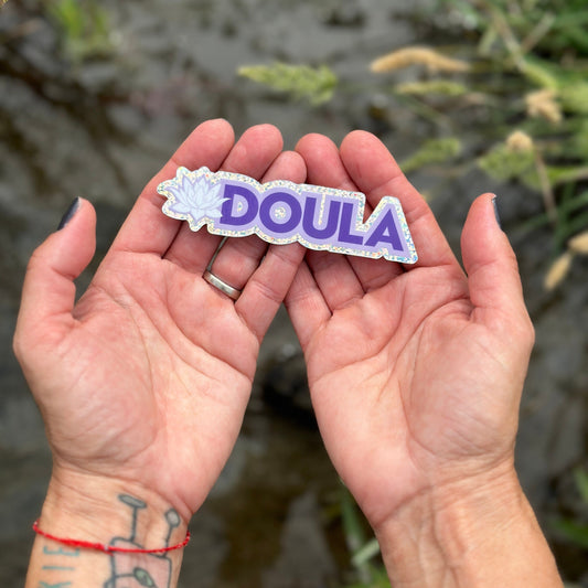 Hand holding a purple 'DOULA' sticker with glittery effect with a blurred natural background.