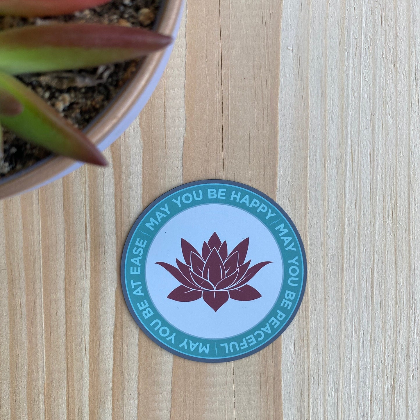 Three inch ultra thin magnet with pink lotus flower design and text, may you be happy may you be peaceful, may you be at ease on a wooden surface with a plant.