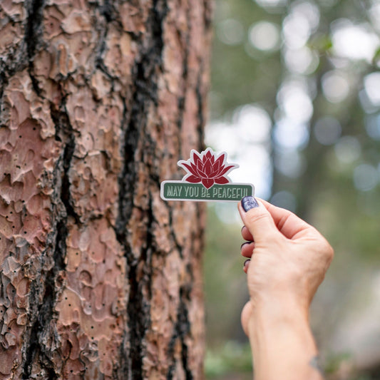 Hand holding a sticker with a red lotus flower design and text, may you be peaceful, against a tree trunk.