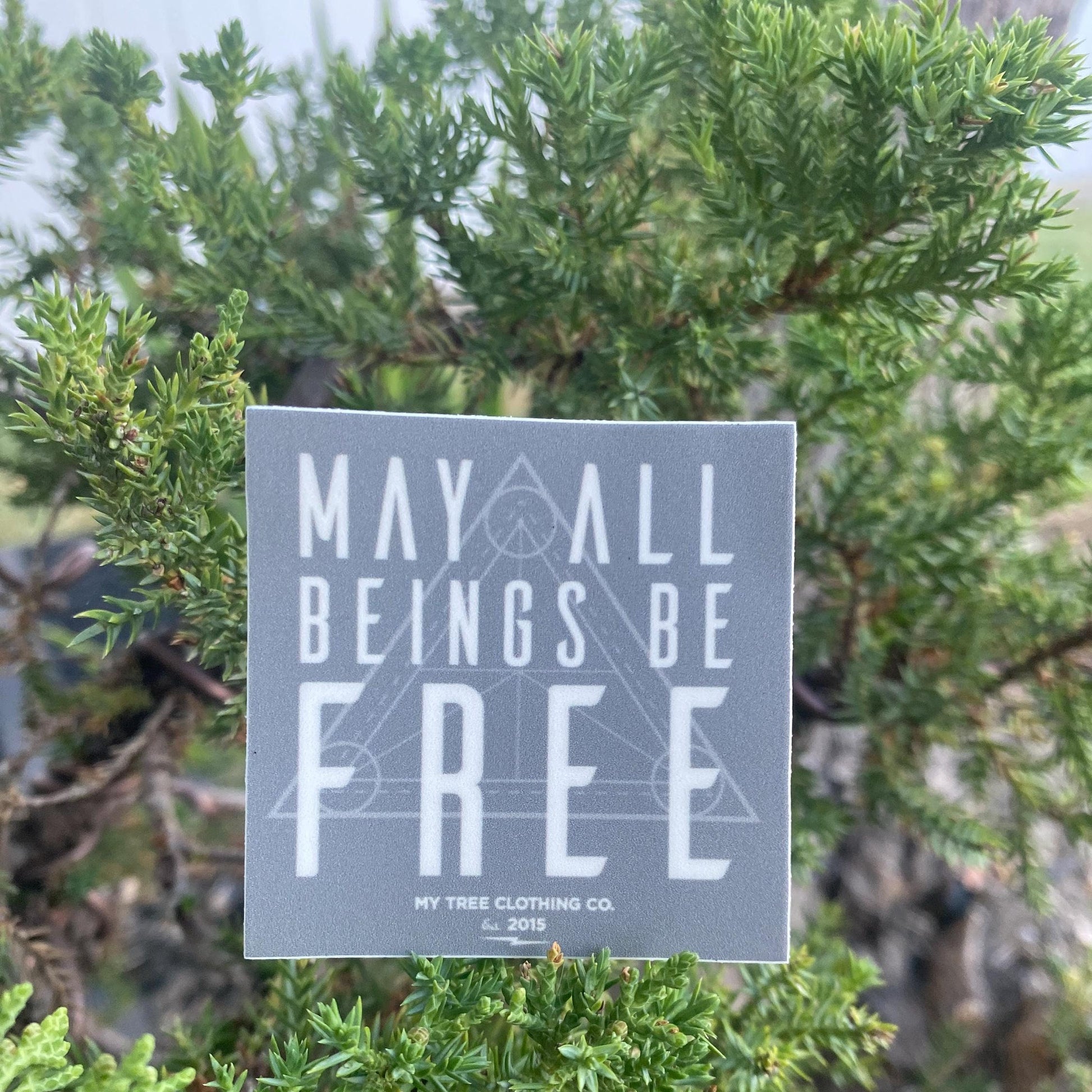 2" square grey sticker with 'May all beings be free' text with sacred geometry design in front of green foliage.