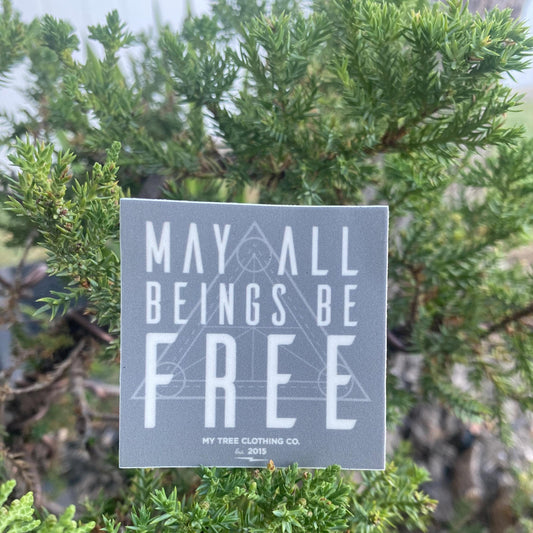 2" square grey sticker with 'May all beings be free' text with sacred geometry design in front of green foliage.