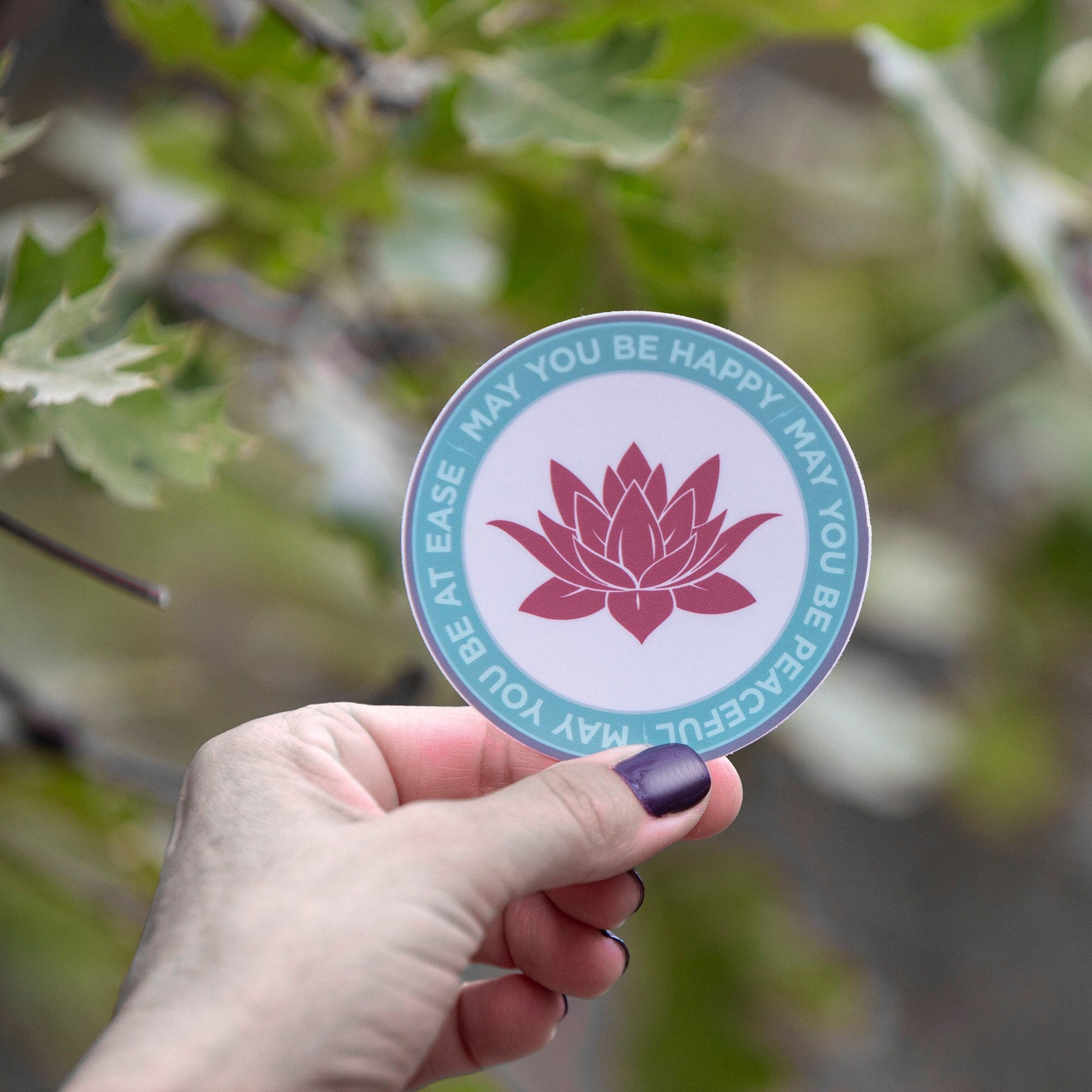 Hand holding a ultra thin magnet with pink lotus flower design and text, may you be happy may you be peaceful, may you be at ease against a blurred natural background.