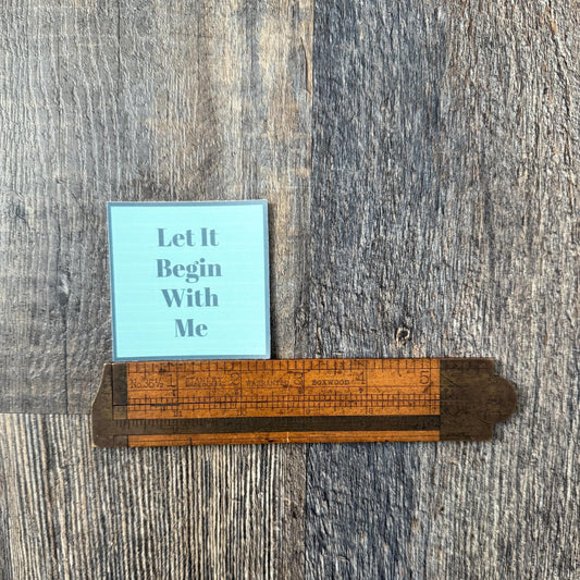 Blue, 2" square sticker with 'Let It Begin With Me' al-anon slogan sticker in blue being measured by an antique wooden ruler.