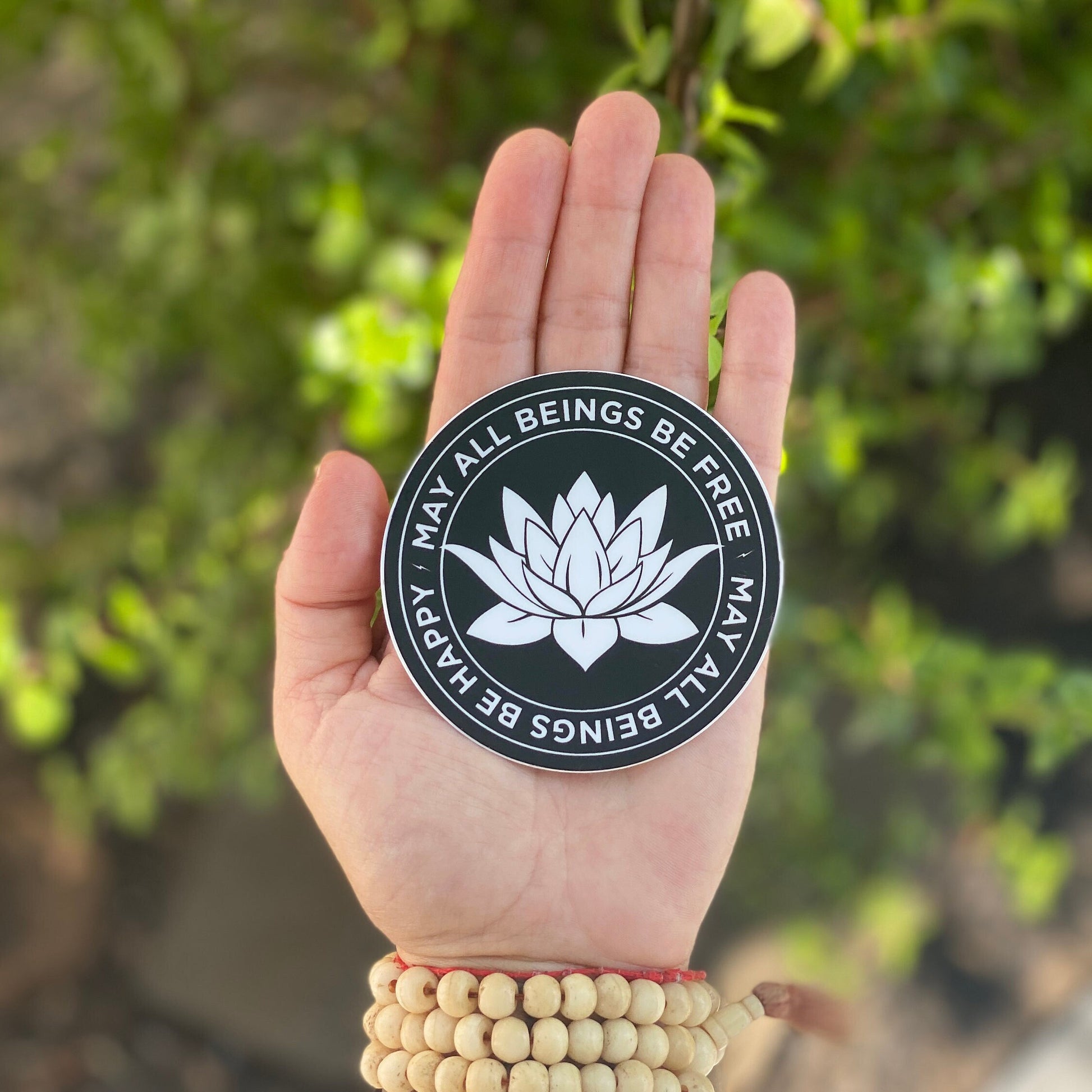 Hand holding a black and white 3" circular sticker with a white lotus flower design and text, may all beings be free may all beings be happy, against a blurred green background.