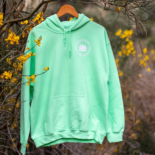 Mint green hoodie with a lotus and the text, may you be happy may you be free may you be peaceful on a wooden hanger against a natural background with yellow flowers.