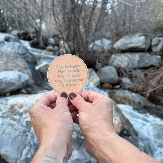 Person holding a circular sticker with text, may i be happy may i be well may i be safe may i be peaceful may i be at ease, against a natural background of rocks and water.