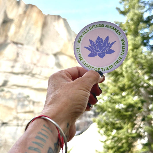 Hand holding a 3" round sticker with a purple lotus flower design and the text, may all beings awaken to the light of their true nature against a natural background.