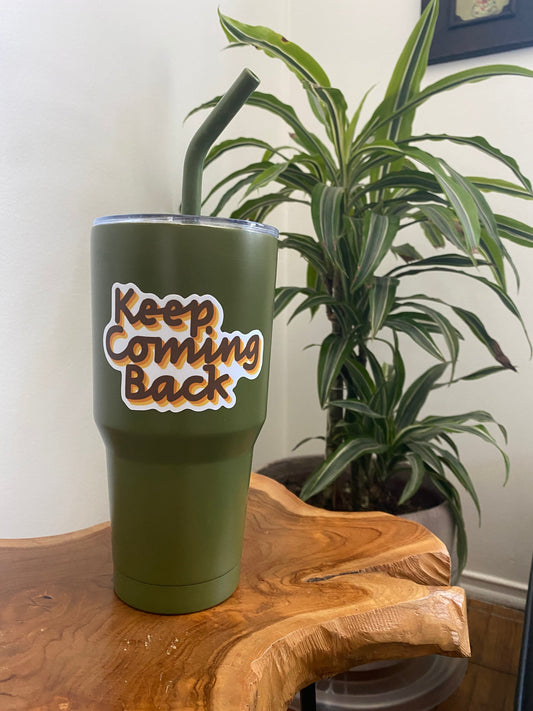 Green tumbler with 'Keep Coming Back' sticker on a wooden surface with a plant in the background.