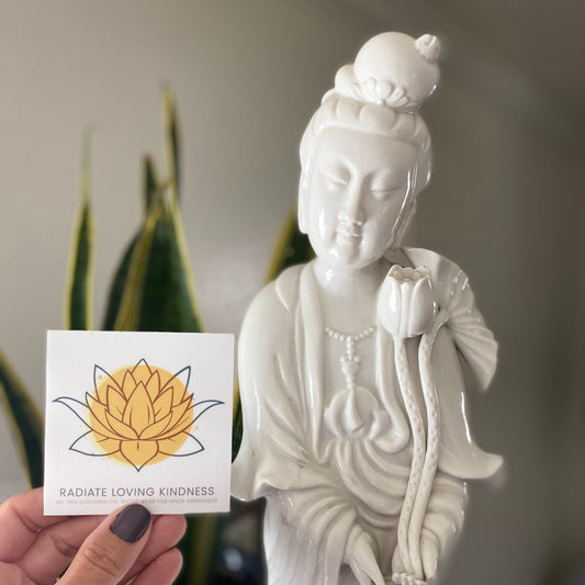 White statue of a seated figure holding a flower with a 'Radiate Loving Kindness' 3" square sticker held in front.