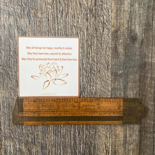 3" square Buddhist sticker with a lotus flower design and text, 'May all beings be happy, healthy and whole. May they have love, warmth and affection. May they be protected from harm and free from fear.' On a wooden surface with a ruler for scale.
