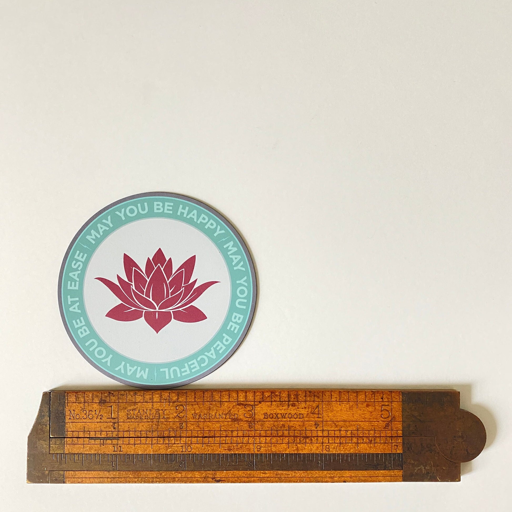 Circular ultra thin magnet with pink lotus flower design and text, may you be happy may you be peaceful, may you be at ease, below a wooden ruler.