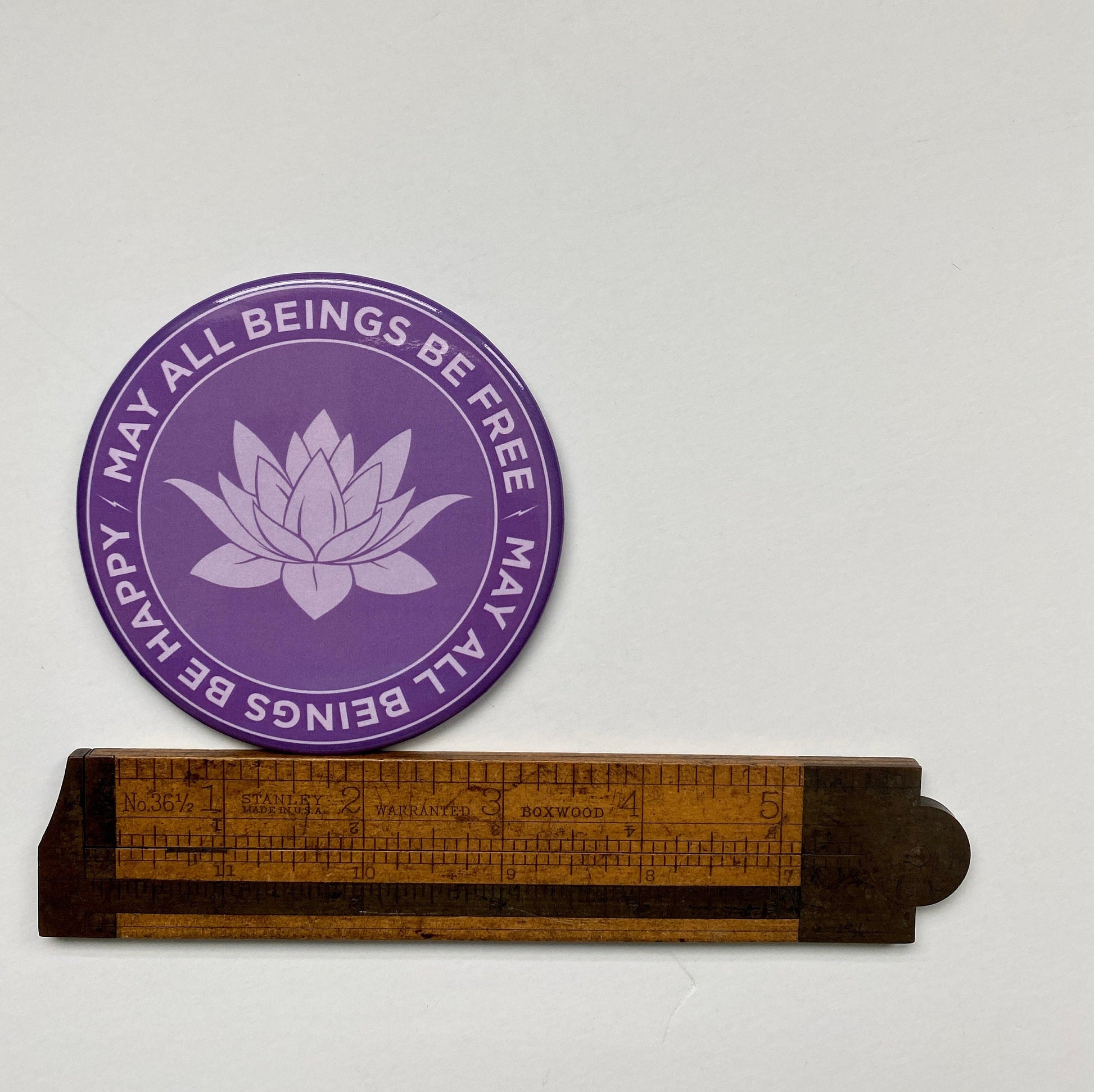 Purple magnet with lotus design and text, may all beings be free may all beings be happy, placed on a white background with a wooden ruler for scale.