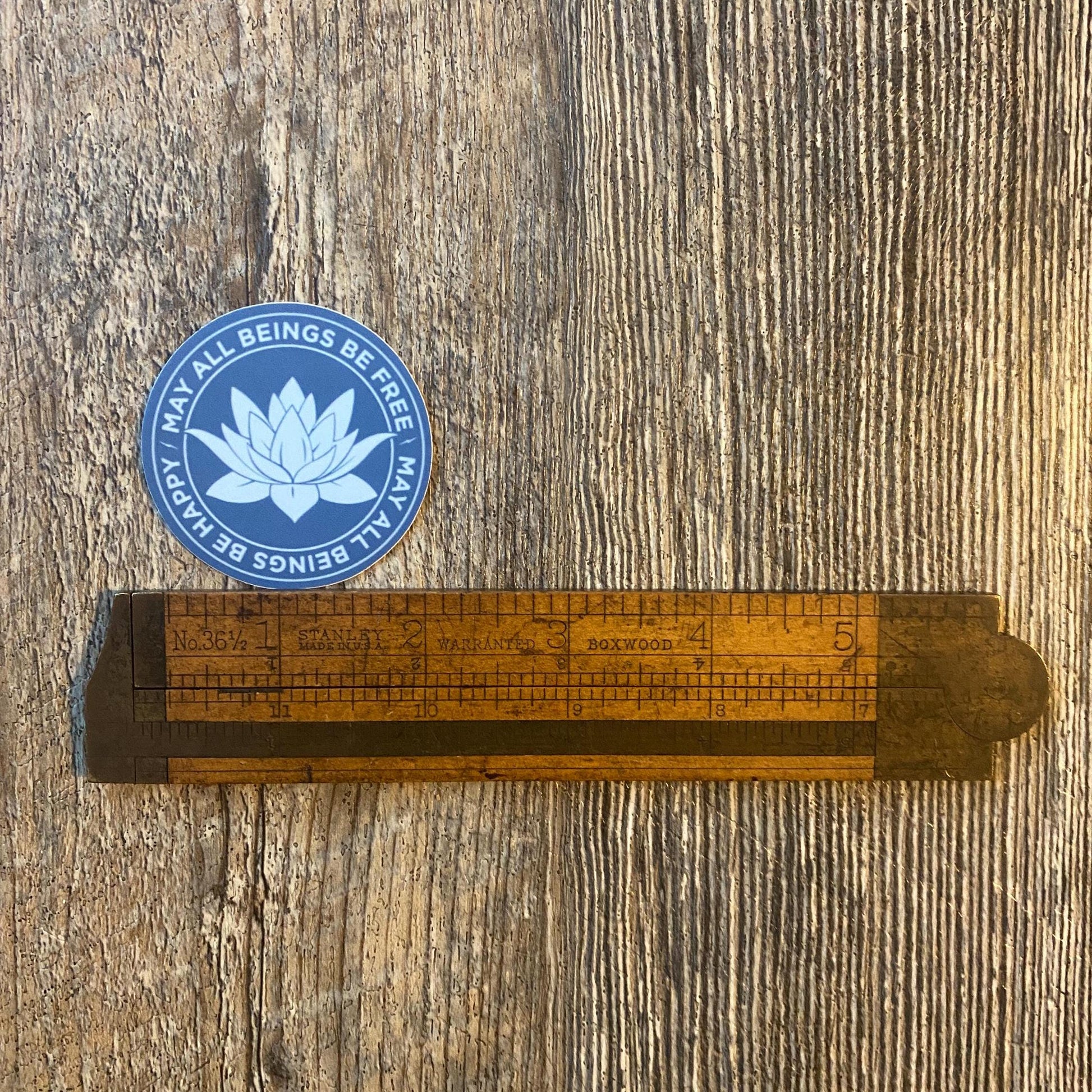 Buddhist meditation sticker in a gorgeous blue with a lotus flower design and the text, 'may all beings be free may all beings be happy' on a wooden surface.