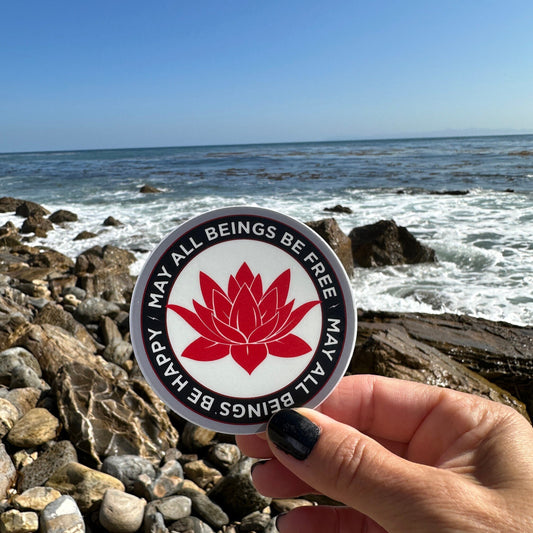 Person holding a circular sticker with a red lotus flower design and text, may all beings be free may all beings be happy, against a rocky beach background.