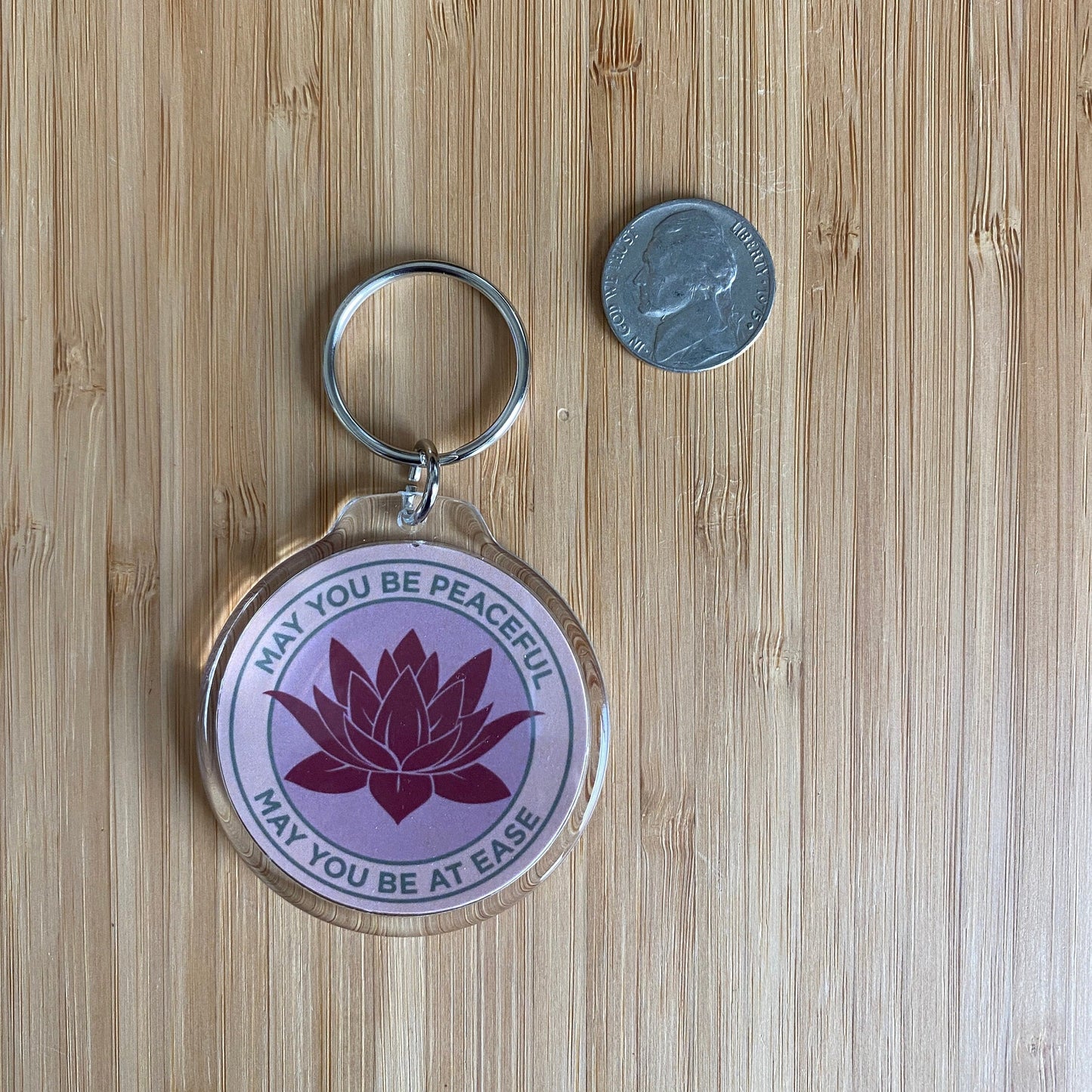 Keychain with lotus flower design and text May You Be Peaceful May You Be At Ease, on a wooden surface next to a coin for size comparison.