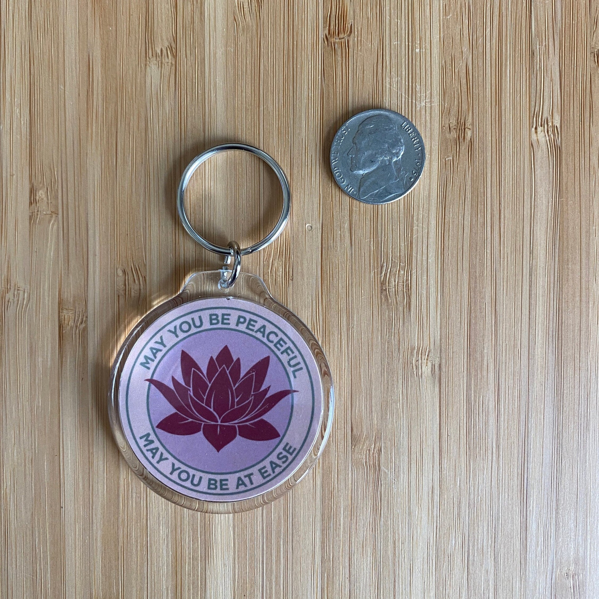 Keychain with lotus flower design and text May You Be Peaceful May You Be At Ease, on a wooden surface next to a coin for size comparison.