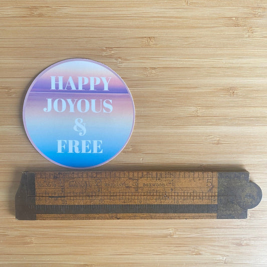 3" round colorful sticker with 'Happy Joyous & Free' text on a wooden surface with a ruler for scale.