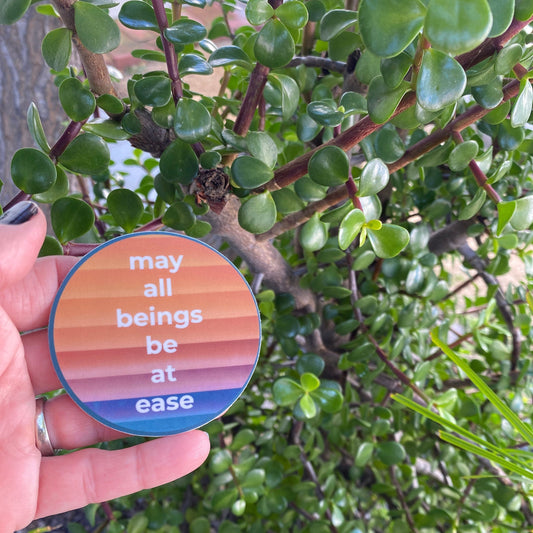 Hand holding a 2.5" colorful rainbow circular sticker with 'may all beings be at ease' text against a green leafy background