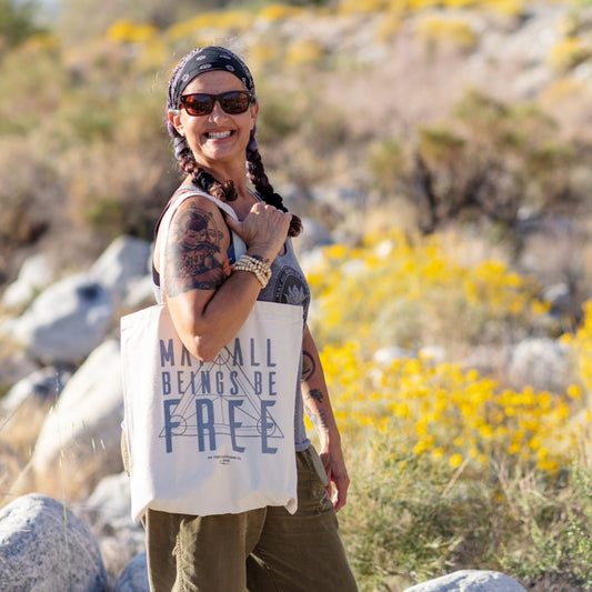 Person holding a tote bag with the text, MAY ALL BEINGS BE FREE, with a sacred geometry design in a desert setting with yellow flowers and rocks.