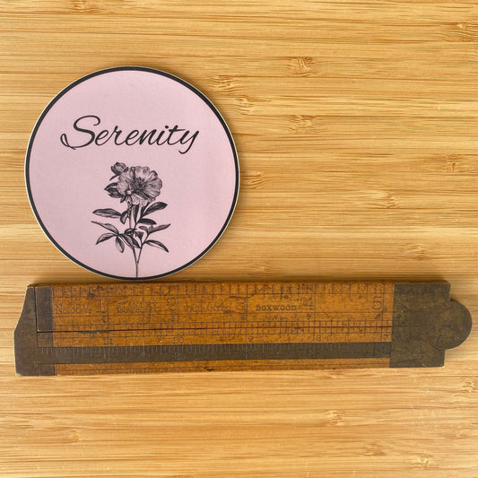 Round pink 12 step recovery sticker with floral design and 'Serenity' text on a wooden surface and a ruler for measurement.