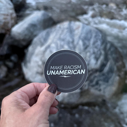 Hand holding a sticker with 'Make Racism Unamerican' text against a rocky background.
