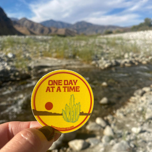 Yellow sticker with 'One Day at a Time' text held over a blurred natural landscape.