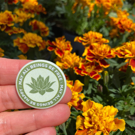 Hand holding a light green 1.25" circular Buddhist pin back button with text and a lotus symbol in front of marigold flowers.