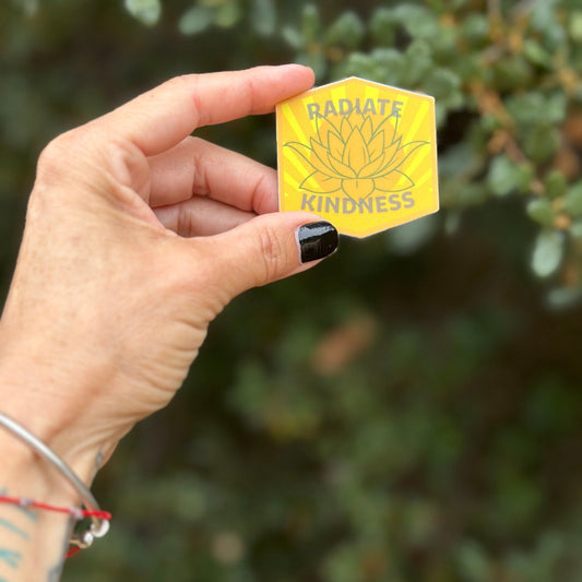 Hand holding a yellow hexagonal sticker with 'Radiate Kindness' text and lotus flower design against a blurred green background.